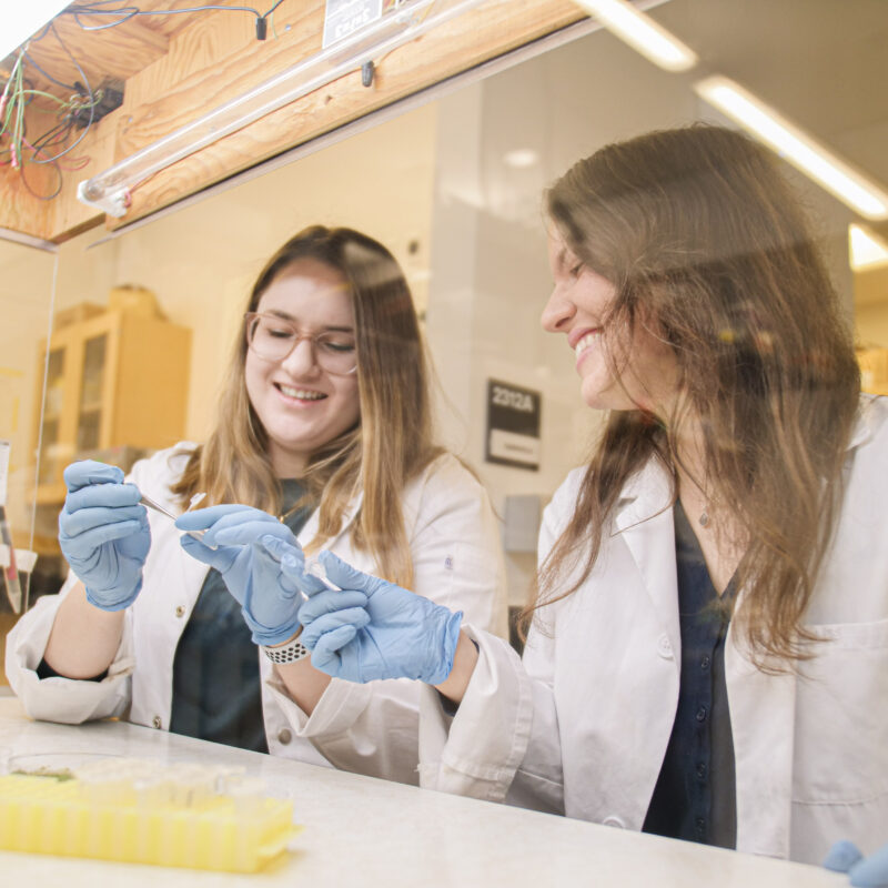 Co-founders Dr. Liz Mahon and Dr. Hila Behar smiling together in a biotech lab, wearing lab coats and holding scientific equipment.