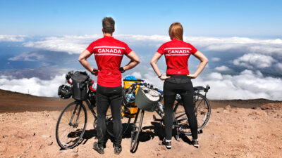 Andreas Bogner and Liuba Mamonova standing side by side on a mountain, facing away from the camera with their bikes next to them, wearing red Canada shirts.